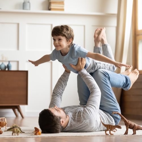 Joyful,Young,Man,Father,Lying,On,Carpet,Floor,,Lifting,Excited Joyful young man father lying on carpet floor, lifting excited happy little child son at home. Full length carefree two generations family having fun, practicing acroyoga in pair in living room.
