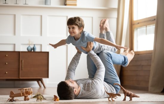 Joyful young man father lying on carpet floor, lifting excited happy little child son at home. Full length carefree two generations family having fun, practicing acroyoga in pair in living room.